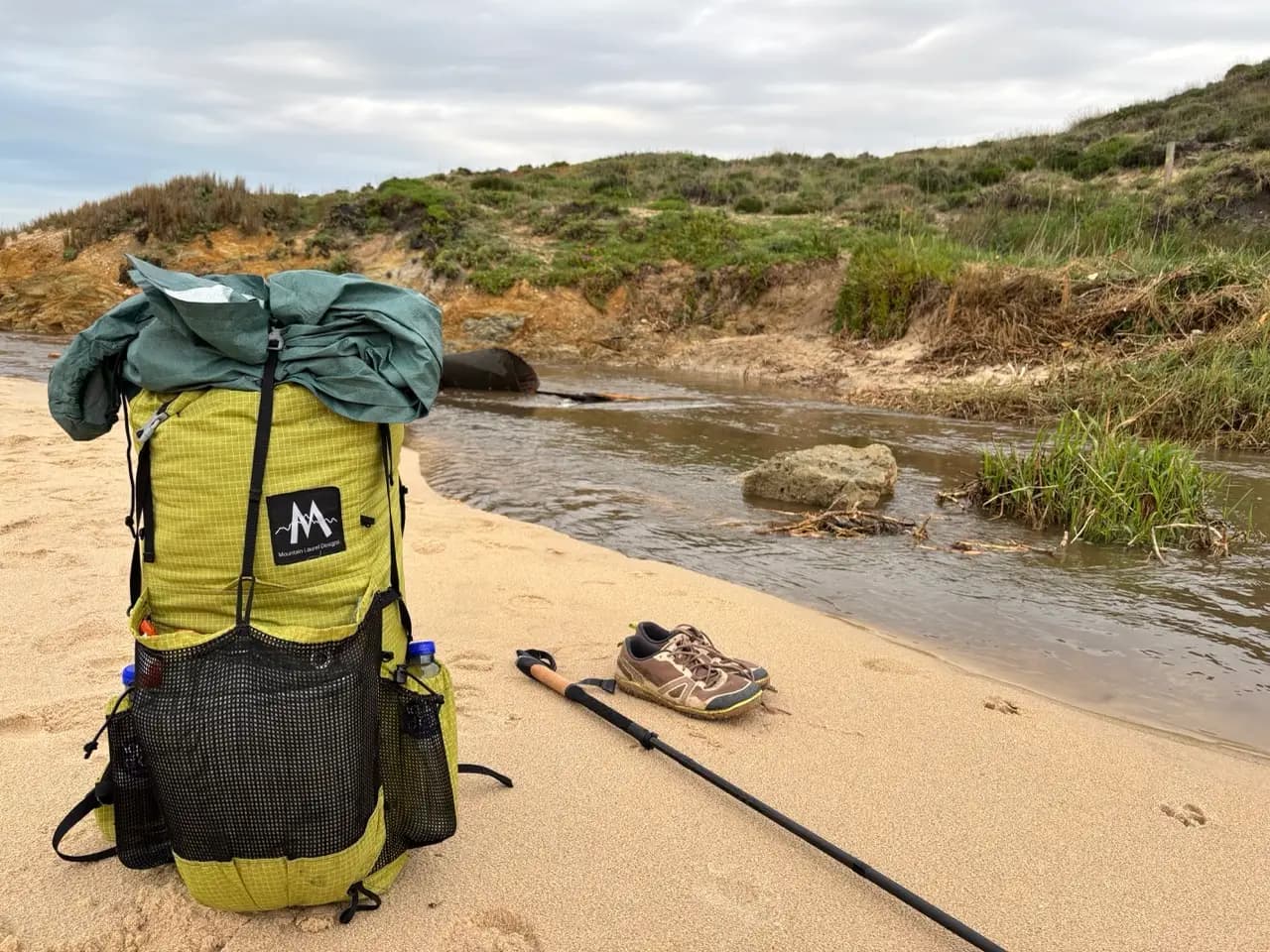 Hiking backpack and trail runners next to a river crossing on the beach